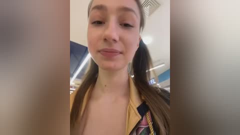 Media: Video of a young woman with light skin and long brown hair in a ponytail, wearing a casual jacket with a colorful plaid pattern. She has a slight smile and is indoors, likely in a public place.