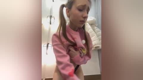 Media: Video of a young, light-skinned girl with pigtails, lifting her pink sweater to reveal her bare chest in a bathroom with a towel rack and shower head.