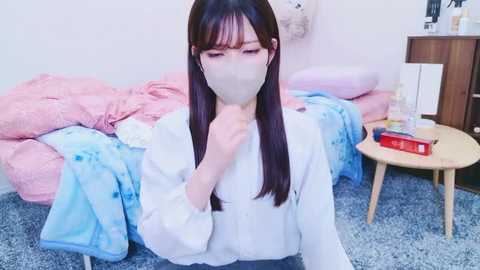 Media: Video of an Asian woman with long black hair, wearing a white shirt, mask, and glasses, sitting on a blue carpet in a room with pink and blue bedding, wooden furniture, and a red book.