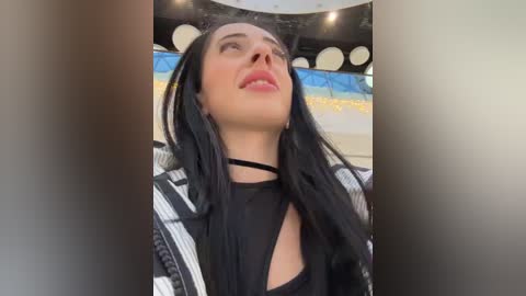 Media: Video of a woman with long black hair and fair skin, wearing a black choker and a black and white striped jacket. She gazes upward, lips slightly parted. Background features modern, circular light fixtures.