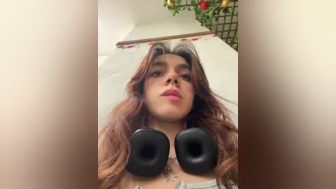 Media: Video of a young woman with long, wavy brown hair, lying on a bed with white sheets, wearing black over-ear headphones, and a slight, contemplative expression.