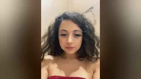 Video of a young woman with curly dark hair, fair skin, and brown eyes, wearing a red strapless top. She stands in a tiled bathroom, looking downcast.