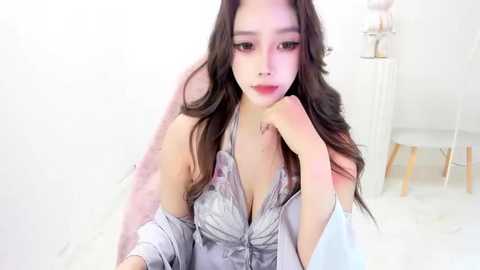 Media: Video of an East Asian woman with long, wavy black hair, fair skin, wearing a revealing, low-cut, silver lace bra with a light pink robe, posing seductively in a minimalist white room.