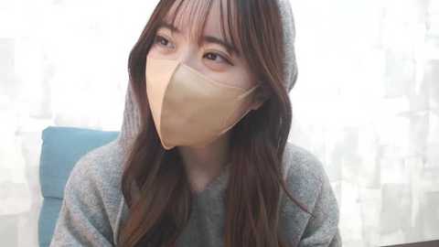 Media: Video of an Asian woman with long brown hair and light skin, wearing a beige face mask, gray hoodie, and sitting in a blue chair.