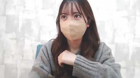 Media: Video of an Asian woman with long black hair, wearing a gray sweater, a beige face mask, and a blue jacket, sitting in front of a white, patterned curtain.