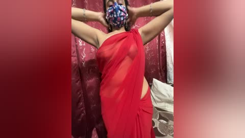 Media: Video of a light-skinned woman with medium-length hair, wearing a red, sheer saree that reveals her breasts and nipples, a mask, and a bracelet. She stands against a red curtain with floral patterns, in a room with a white bed and a pile of clothes.