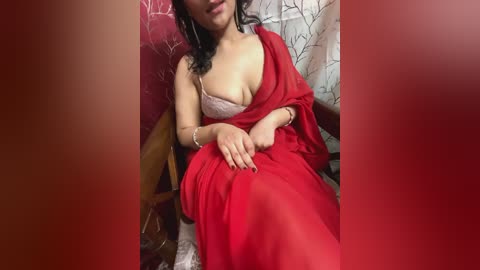 Media: Video of a South Asian woman with medium skin tone, wearing a red saree, revealing a pink bra. She sits on a wooden chair with floral-patterned curtains in the background.