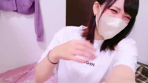 Media: A video of a young Asian woman with shoulder-length black hair, wearing a white T-shirt, a white mask, and a black bracelet. She is indoors, with a purple towel hanging on a wall and a checkered blanket visible.