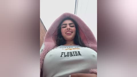 Media: Video of a young woman with long dark hair, wearing a white \"Florida\" t-shirt, covered by a pink hooded towel. She smiles, revealing a slight cleavage. Background shows a blurred indoor setting.