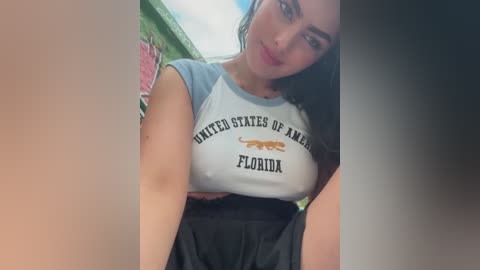 Media: Video of a young woman with long dark hair, wearing a tight, white and blue \"United States of America\" t-shirt, showing cleavage, and black shorts, posing indoors.