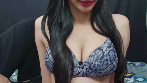 Media: Video of a light-skinned woman with long, wavy black hair, wearing a black and gray patterned bra, sitting against a dark background, lips painted red.