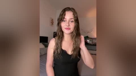 Video of a young Caucasian woman with wavy brown hair, wearing a black tank top, standing in a modern bedroom with gray bedding and string lights.