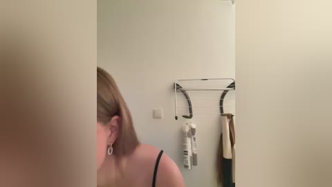 Media: A video of a woman with blonde hair in a black tank top, partially visible, standing in a minimalist bathroom with a white wall and metallic towel rack holding a bottle.