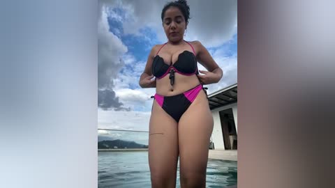 Media: Video of a Latina woman with medium skin tone, wearing a black and pink bikini, standing by a pool under a cloudy sky with mountains in the background.