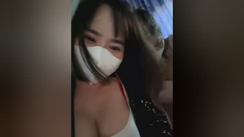 Media: Video of an Asian woman with short, dark hair and a white mask covering her mouth. She is wearing a red and white tank top, and the background features a blue curtain and a shiny, reflective surface.