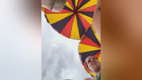 Media: A video of a man with light skin and short hair, wearing a green shirt, holding a vibrant umbrella with a red, yellow, and black striped pattern, against a cloudy sky background.