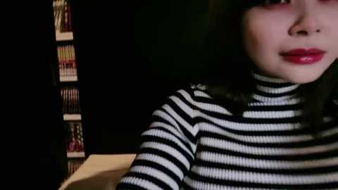 Media: Video of a young woman with fair skin and dark hair, wearing a black and white striped turtleneck sweater, against a dark, bookshelf-lined background.