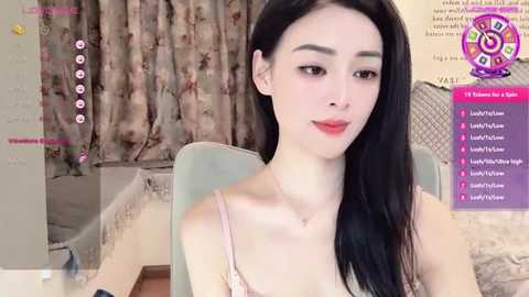 Media: A video of an Asian woman with long black hair, fair skin, and a slender physique, wearing a light pink camisole. She sits indoors with floral curtains in the background.