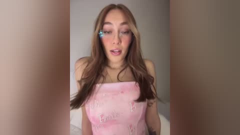 Media: Video of a young woman with long, light brown hair, wearing a pink bandeau top with \"Barbie\" written on it. She has a light skin tone, blue hair clips, and is looking directly at the camera.