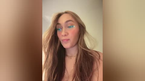 Media: Video of a young woman with long, light brown hair, wearing turquoise eyeshadow, a gold necklace, and a subtle, confident smile, set against a neutral beige background.