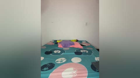 Media: A video of a minimalist, brightly colored children's bedroom with a single bed covered in a turquoise bedspread featuring pink, white, and blue circular patterns. The room has white walls and a small, plain window.