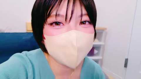 Media: Video of a young Asian woman with short black hair, wearing a light blue robe and a beige surgical mask, in a sterile medical setting with white walls and a blue chair.