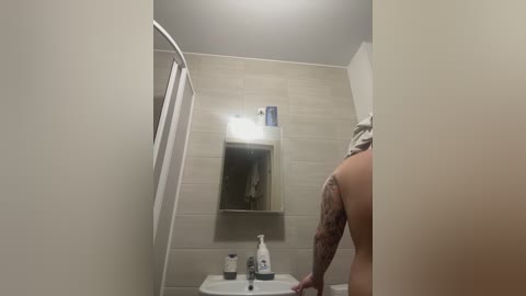 Media: Video of a small, beige-tiled bathroom with a white sink, mirror, and towel rack. A person with a tattooed arm stands in the background, partially visible.