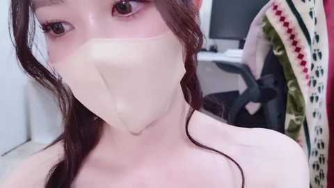 Media: Video of a young Asian woman with fair skin, wearing a surgical mask, brown hair, and a white top, in a room with a desk, chair, and a colorful patterned cloth.