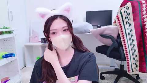 Media: Video of a young Asian woman with long brown hair, wearing a black T-shirt, white cat ears headband, and face mask, sitting on a carpeted floor in a room with white walls, shelves, and a desk.