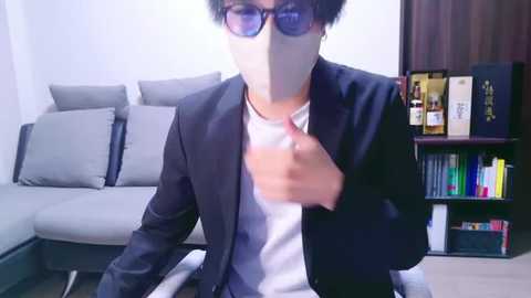 Media: Video of a man with short black hair, glasses, and a white mask, wearing a black suit and white shirt, standing in a modern living room with gray couches, wooden bookshelves, and bottles of liquor.