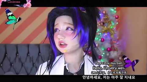 Media: Video of a fair-skinned woman with long black hair and blue highlights, wearing a white jacket, in a colorful room with a Christmas tree.