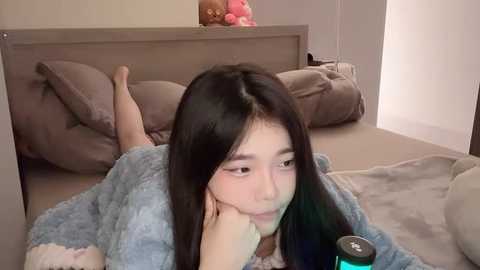 Media: Video of an East Asian woman with long black hair, lying on her stomach in a plush robe, looking contemplative. A stuffed toy and a green device are visible in the background.