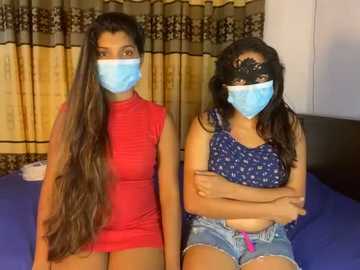 Media: Video of two South Asian women with long, wavy hair, wearing blue surgical masks, red and blue patterned tops, and denim shorts, sitting on a bed with dark curtains in the background.