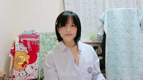 Media: Video of a young Asian woman with short black hair and a light blue shirt, sitting in a cluttered room with colorful, patterned bedding and a white curtain with polka dots in the background.