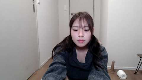 Media: A video of a young Asian woman with long black hair, wearing a dark sweater, sitting on a wooden floor in a minimalistic hallway, with a roll of toilet paper beside her.