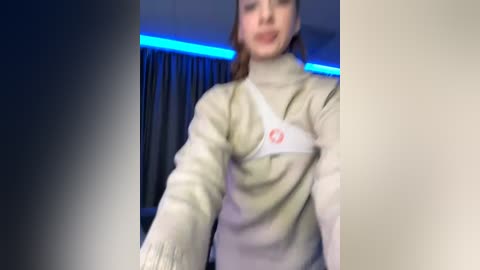 Media: Video of a woman in beige sweater with a white bandage on her chest, standing in a dimly lit room with blue LED strip lighting, curtains in the background.