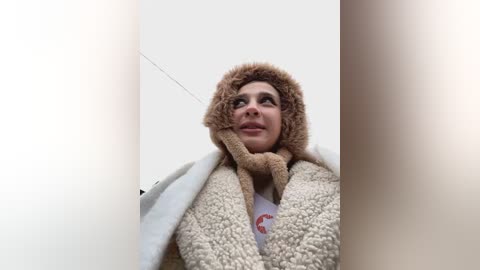 Media: Video of a woman with fair skin, wearing a fuzzy, brown, animal-themed hood, and a white coat, looking upwards into a narrow, beige-tiled space.