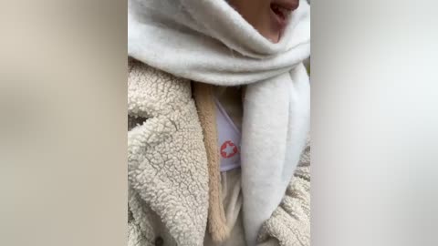 Media: A video of a person's neck and upper chest, wearing a beige, fluffy robe and a white scarf, partially revealing a red cross emblem on a white shirt.