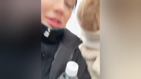 Media: A blurred video of a close-up scene showing a person in a black jacket and beige scarf, with a light-skinned person in a striped sweater in the background.