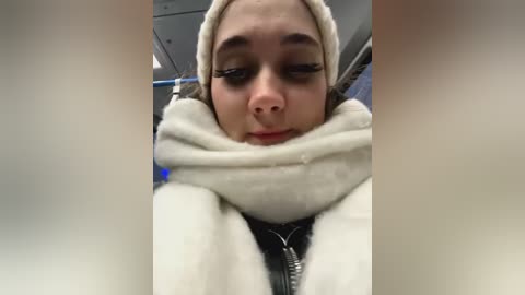 Media: A close-up video of a woman with light skin, wearing a beige scarf and a black jacket, with long eyelashes and a neutral expression, taken in an indoor setting.