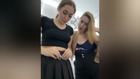 Media: Video of two young women in a bathroom. One wears a black crop top and skirt, adjusting her belt. The other, in a navy shirt and high-waisted pants, stands behind her. Background shows a white tiled wall and a blurred person in the foreground.