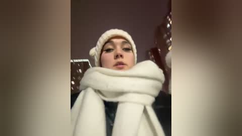 Media: A video of a young woman with pale skin and dark hair, bundled in a thick white scarf and knit hat, standing indoors with blurred background.