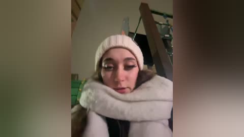 Media: Video of a young woman with fair skin, wearing a beige knitted beanie and a white fluffy jacket, hugging herself, indoors, blurry background.