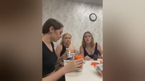 Media: Video of four women, three with fair skin, one with light brown skin, seated at a table, drinking from orange cups, in a room with beige wallpaper and a wall clock.