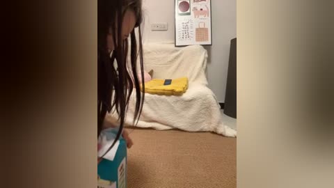 Media: Video of a person with long dreadlocks, wearing a yellow towel, standing on a beige carpet in a room with white walls and a white blanket-covered chair.