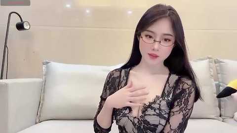 Media: Video of a young Asian woman with fair skin, long black hair, and glasses, wearing a black lace bodysuit, sitting on a white sofa, indoors.