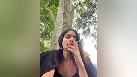 Media: Video of a young woman with long black hair, wearing a black cape, smoking a cigarette outdoors in a forest with tall trees. Her eyes are closed, and her expression is contemplative.