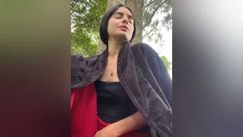 Media: A video of a woman with short black hair, wearing a black top and red skirt, under a dark cape, outdoors against a backdrop of green trees and sky.