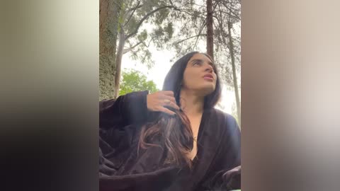 Media: Video of a woman with long dark hair, wearing a black robe, standing in a forest, looking up at the sky, with trees and greenery in the background.