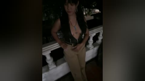 Media: A dimly lit video of a woman with short, dark hair wearing a black leather vest and beige pants, standing on a balcony railing, looking downward.